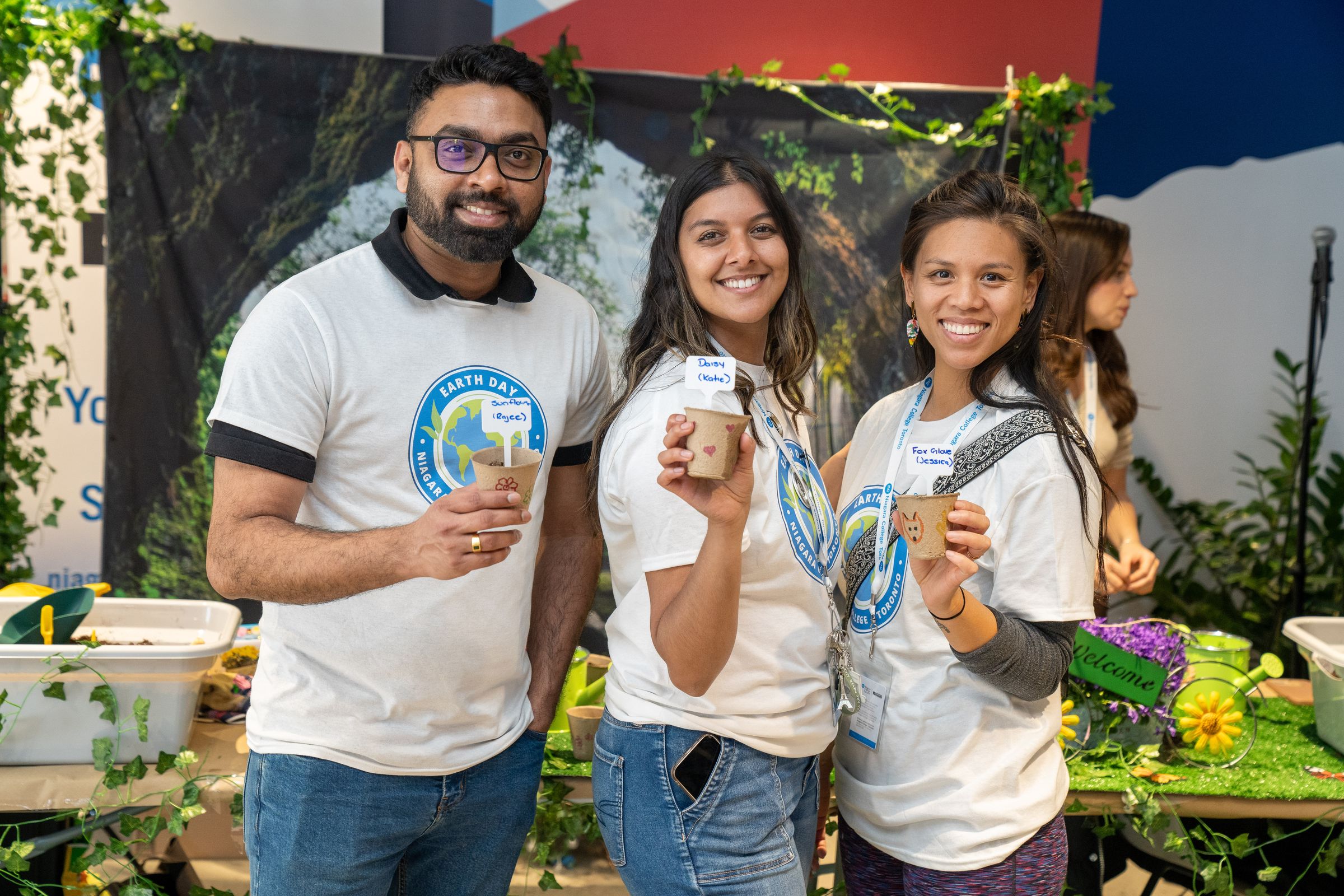 Three people smiling, holding small plant pots, in a decorated booth with greenery. They are wearing matching Earth Day t-shirts.