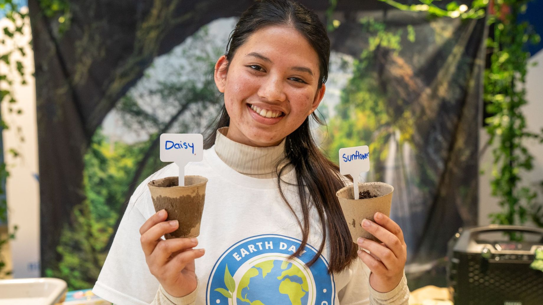 A volunteer at an Earth Day event holds two small biodegradable pots labeled "Daisy" and "Sunflower," each containing soil for planting. The person is wearing a white turtleneck sweater and an Earth Day Niagara Toronto t-shirt. The background features a nature-themed setup with green vines, trees, and a forest backdrop, creating an eco-friendly atmosphere. The volunteer has a warm smile, conveying enthusiasm for environmental initiatives.