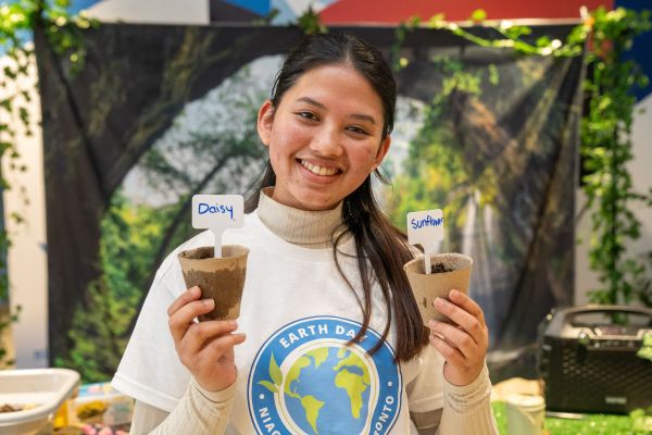 A volunteer at an Earth Day event holds two small biodegradable pots labeled "Daisy" and "Sunflower," each containing soil for planting. The person is wearing a white turtleneck sweater and an Earth Day Niagara Toronto t-shirt. The background features a nature-themed setup with green vines, trees, and a forest backdrop, creating an eco-friendly atmosphere. The volunteer has a warm smile, conveying enthusiasm for environmental initiatives.