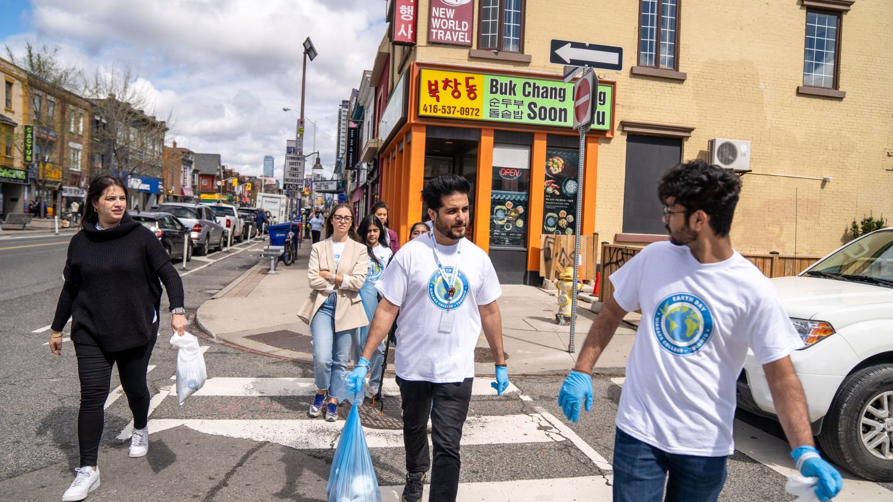 People wearing gloves and holding garbage bags walk across a crosswalk, participating in a street cleanup event in an urban area with various shops and buildings in the background.