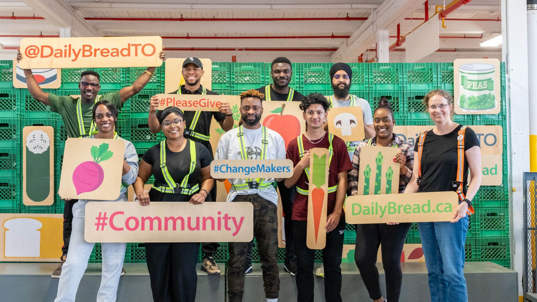 A group of volunteers at a food bank pose for a photo while holding signs with messages such as "@DailyBreadTO," "#Community," "#PleaseGive," "#ChangeMakers," and "DailyBread.ca." Some hold illustrations of vegetables like beets, carrots, mushrooms, and asparagus. They are wearing high-visibility vests or safety harnesses, standing in front of green crates filled with food supplies. The setting is an indoor warehouse with industrial lighting, red ceiling pipes, and organized food storage. The group appears cheerful and engaged in their volunteering efforts.