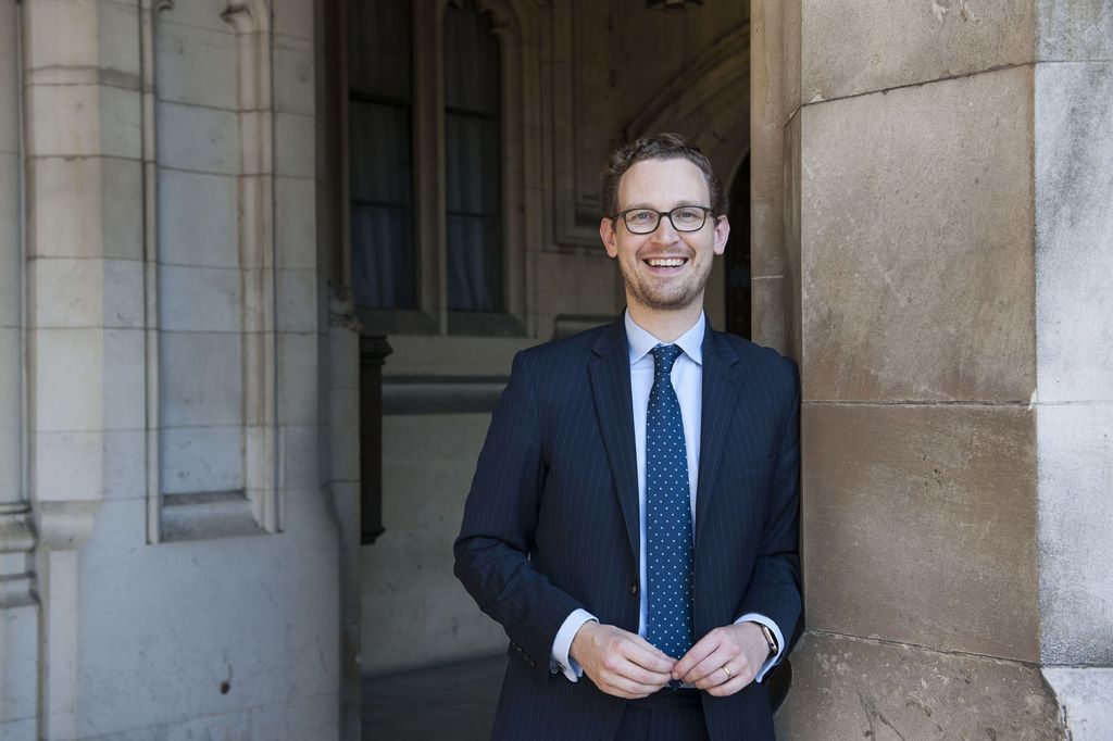 Smiling person in navy pinstripe suit and glasses standing by stone archway of historic building.