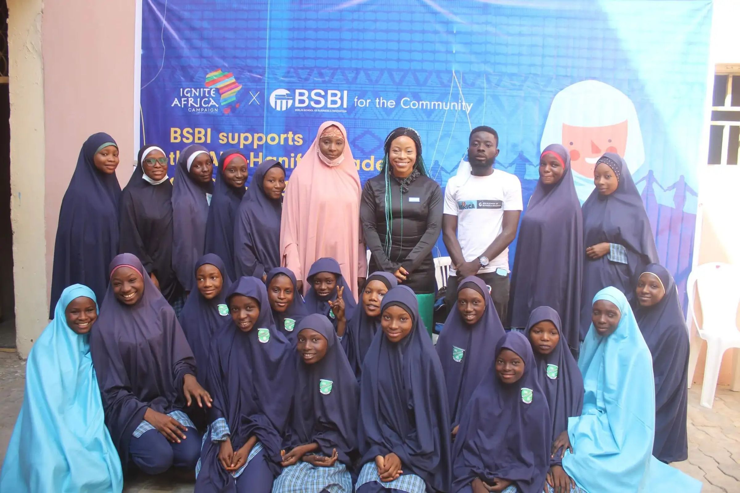A group of students and community members pose for a photo in front of a banner displaying the logos of "Ignite Africa Campaign" and "BSBI for the Community." The banner indicates support for an educational initiative. Many of the students are wearing navy blue and light blue hijabs, while some adults in the group are dressed in different attire, including a pink hijab and a black jacket. The setting is outdoors with a tiled floor and a light-colored building wall in the background. The group appears engaged and supportive of the community initiative.