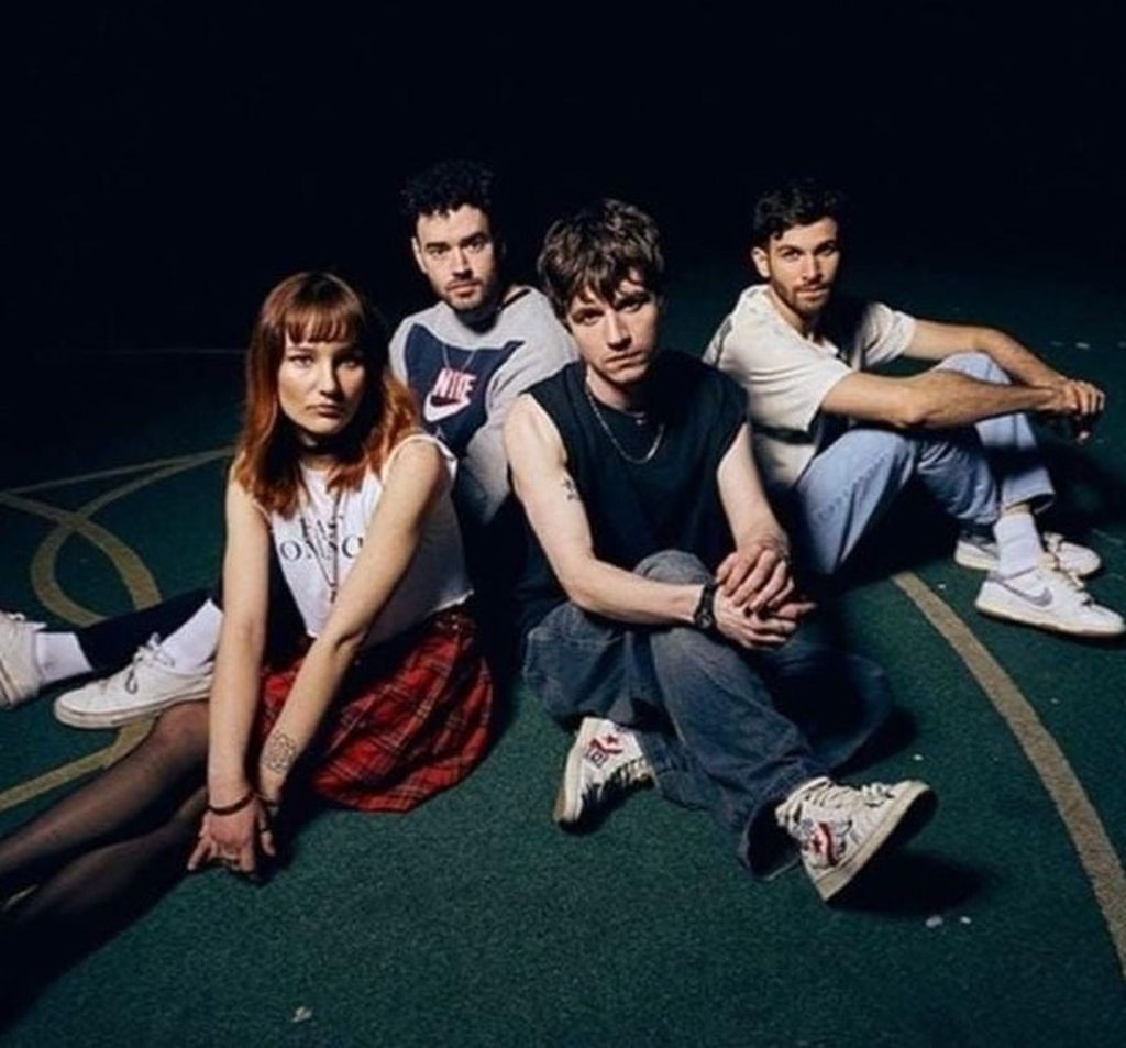 Four young people seated on a dark green floor in casual attire, posing for a band or group photo.