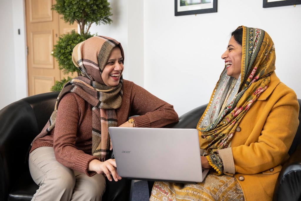 Two women in hijabs sitting in black chairs, smiling and looking at each other. One wears a brown scarf and holds a laptop, while the other is in a yellow outfit with a patterned scarf. A potted plant and framed pictures are in the background.