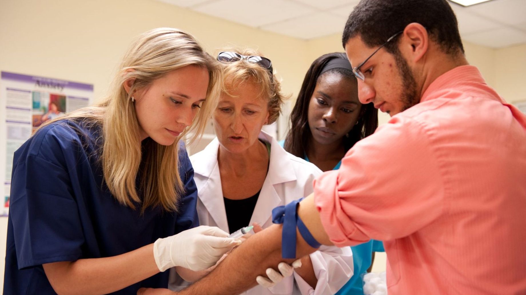 Medical instructor in white coat demonstrating injection technique to three healthcare students in a training lab.