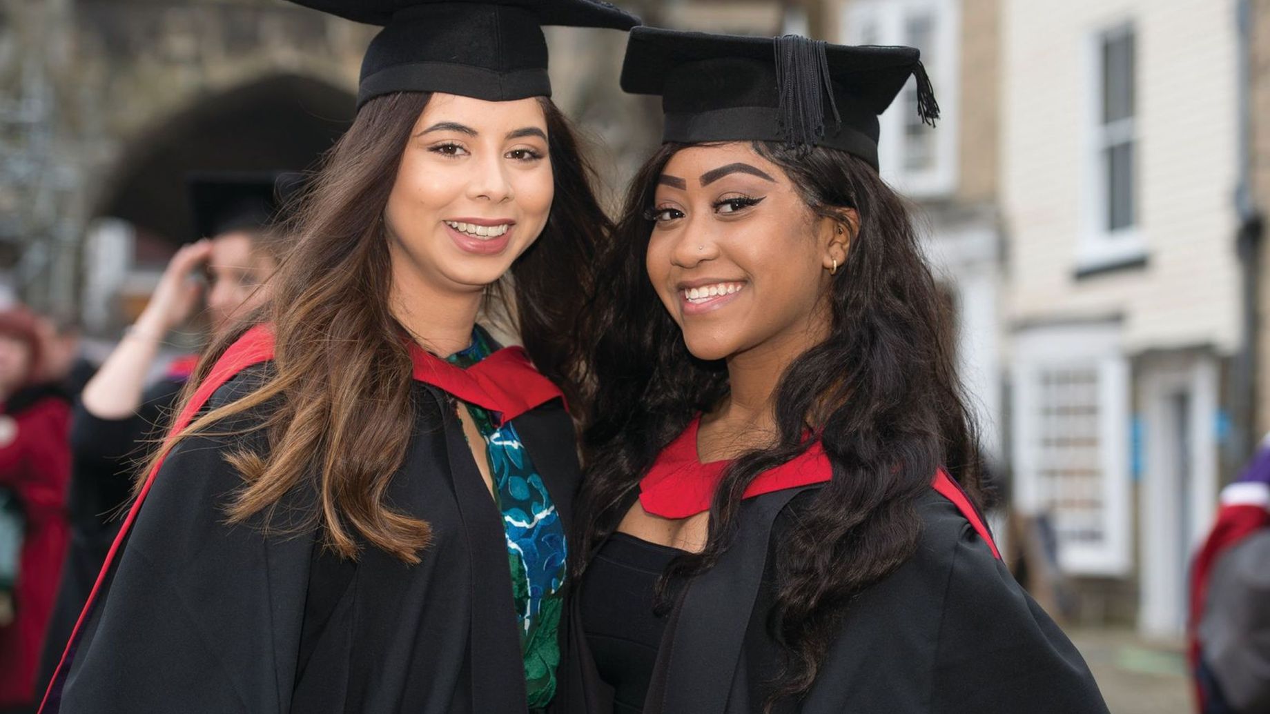 Two graduates in black caps and gowns with red stoles smiling together outdoors during a graduation ceremony.