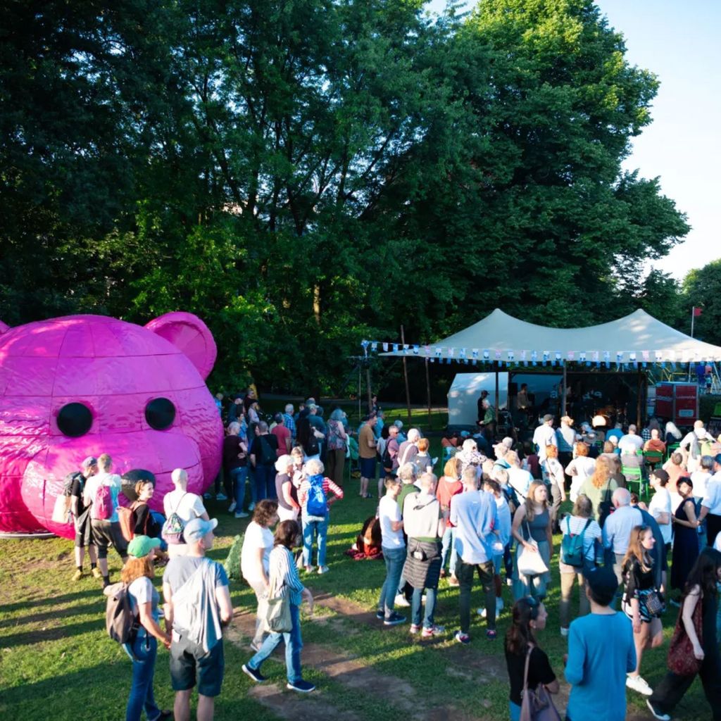 A large crowd gathers around a giant pink bear under a blue sky in a park. A white tent is set up in the background among trees. People are walking and chatting, enjoying the outdoor event.