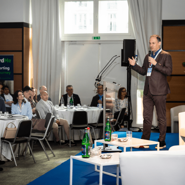 A speaker addresses an audience seated at round tables in a bright room with large windows and a clear podium.