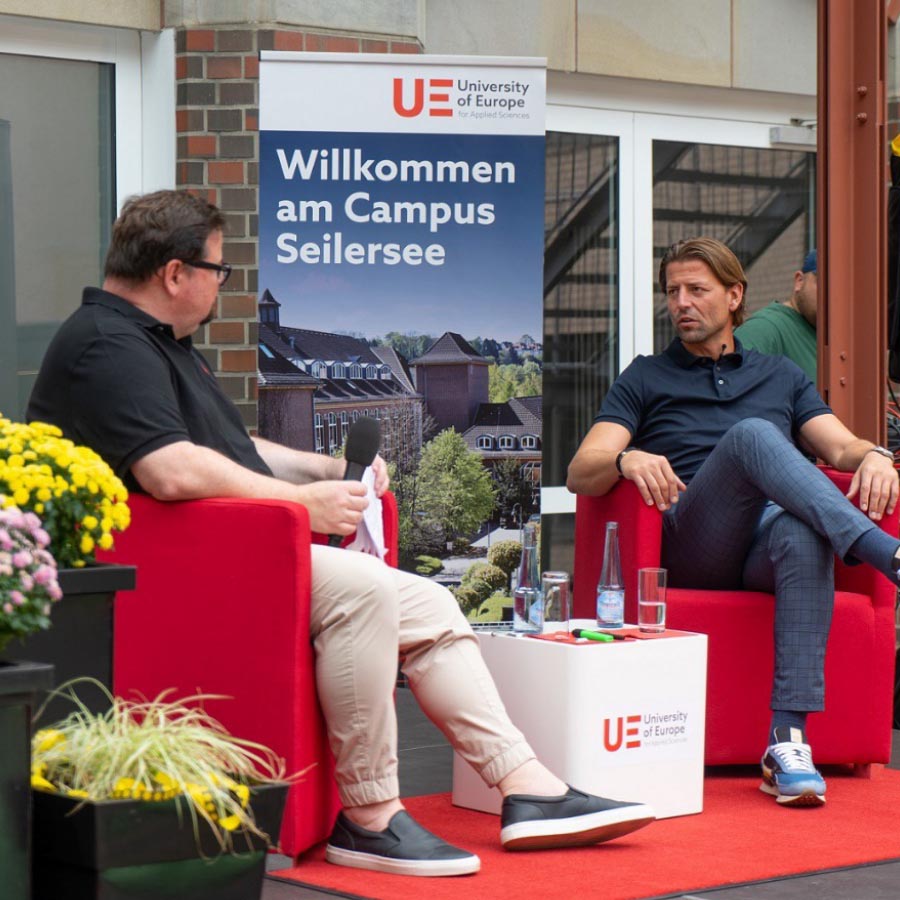 Two people sitting on armchairs talking, either side of a UE backdrop.