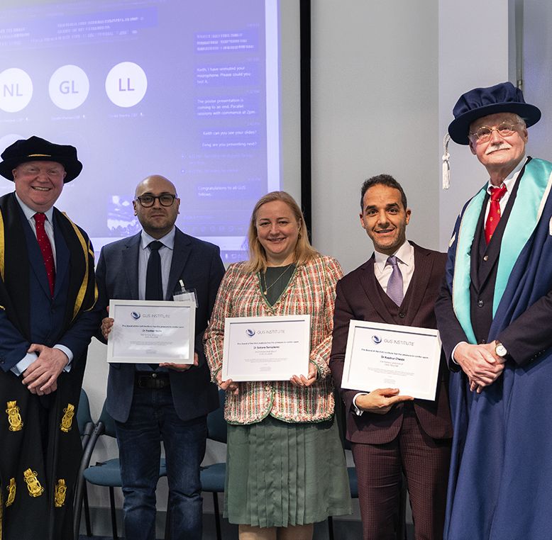 Three graduates holding certificates standing with two academic officials in ceremonial robes and caps at a graduation ceremony.
