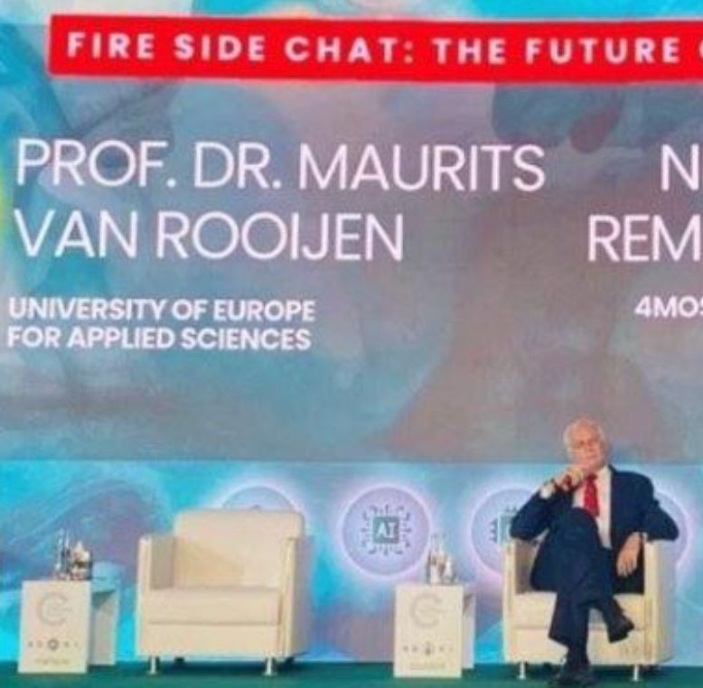 Fireside chat stage with "PROF. DR. MAURITS VAN ROOIJEN" on screen and a person seated in a white chair.