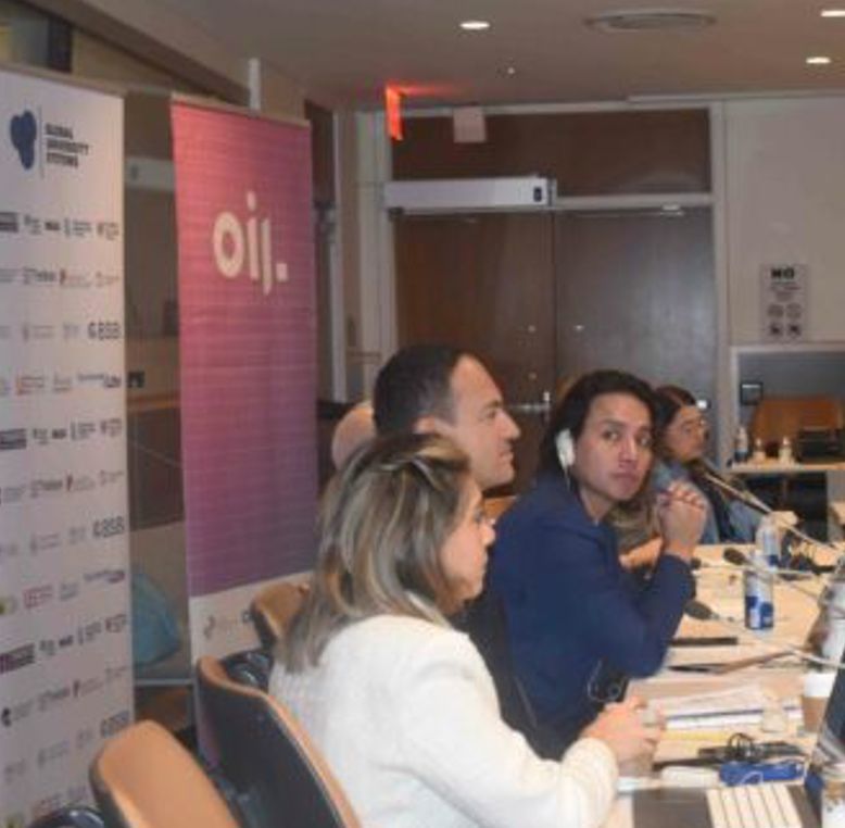 A group of people sit at a table in a conference room, listening to a presentation. Banners with logos are visible in the background.