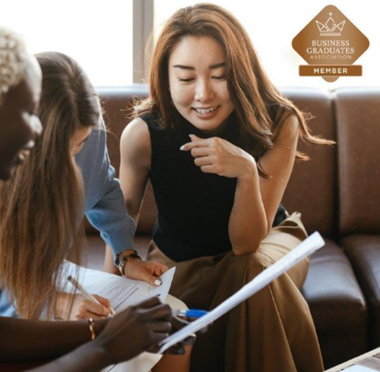 Business meeting with two women discussing documents, Business Graduates Association logo in corner.