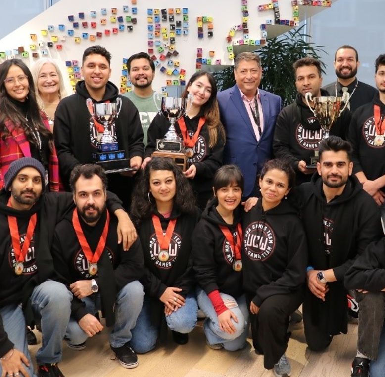 A diverse group, many in University Canada West MBA Games hoodies, pose with trophies and medals against a colourful wall.