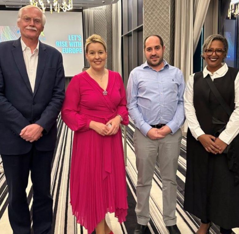 Four professionally dressed individuals stand together in a modern event space with a screen in the background.