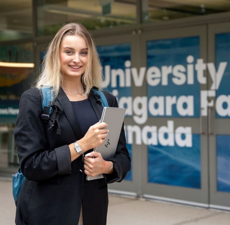A University of Niagara Falls student standing outside the campus, holding a laptop and smiling at the camera.