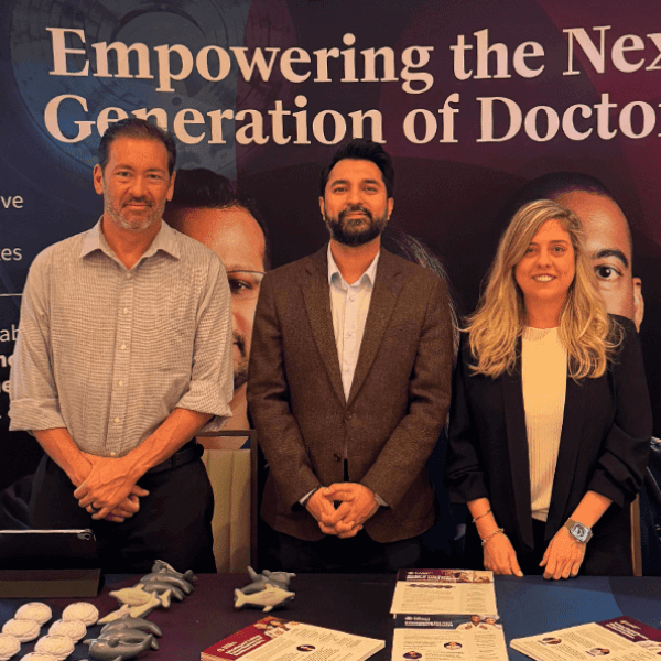Two men and one woman stand in front of a banner that reads 'Empowering the Next Generation of Doctors', with brochures and model airplanes on a table.