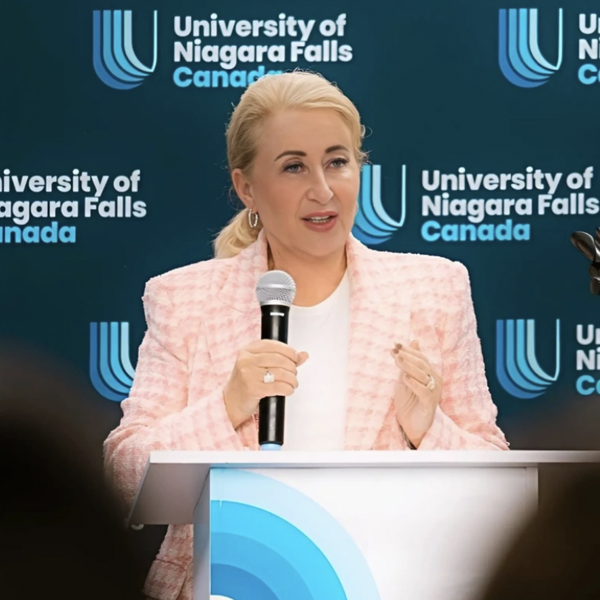 A speaker in a light pink jacket stands at a podium with a microphone, in front of a backdrop featuring the University of Niagara Falls Canada logo.