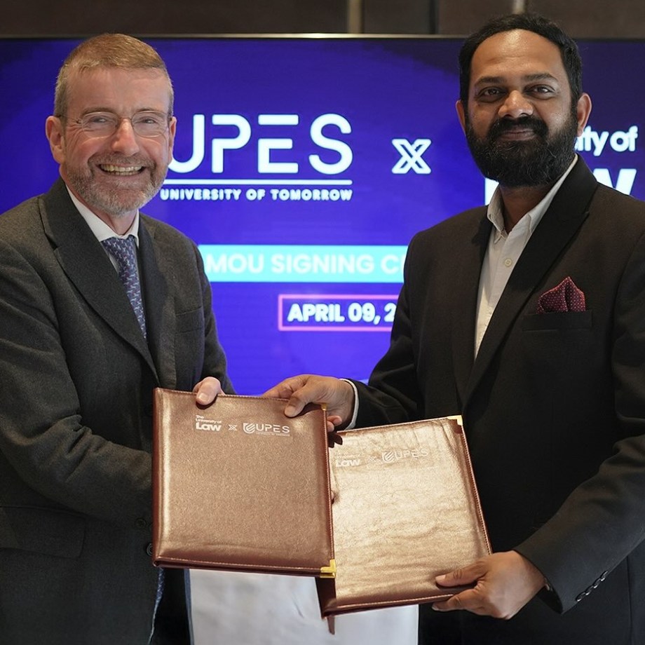 Two individuals in formal attire shaking hands and holding folders during an MoU signing ceremony. The background screen reads 'UPES UNIVERSITY OF TOMORROW' and 'MOU SIGNING CEREMONY APRIL 09. The folders display logos of 'School of Law' and 'UPES.