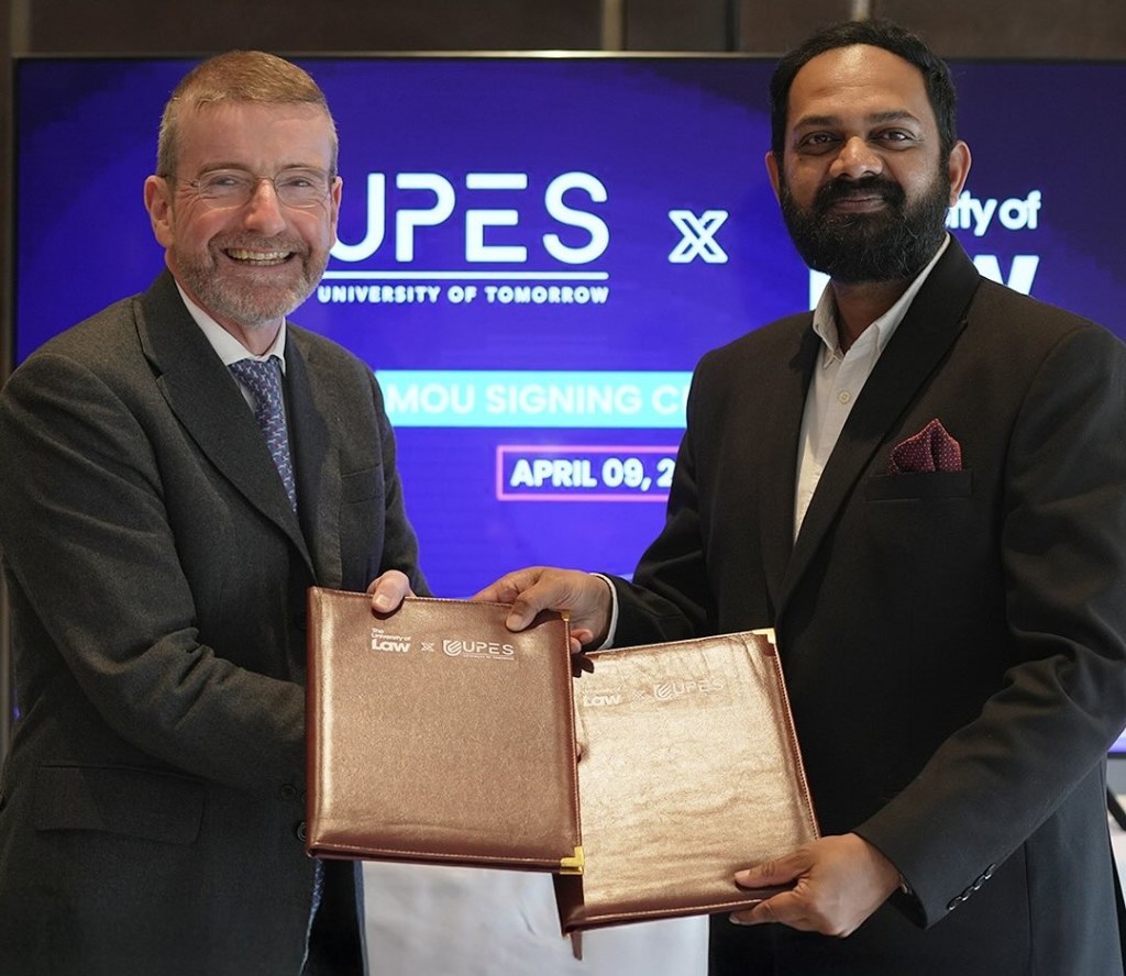 Two individuals in formal attire shaking hands and holding folders during an MoU signing ceremony. The background screen reads 'UPES UNIVERSITY OF TOMORROW' and 'MOU SIGNING CEREMONY APRIL 09. The folders display logos of 'School of Law' and 'UPES.