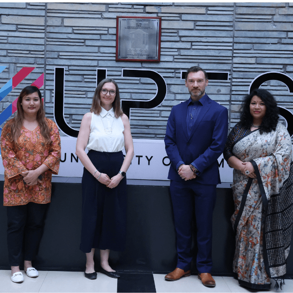 Four individuals in formal attire standing in front of a wall displaying the text 'UPES'. Two members are from UPES and two from the University of Edinburgh.