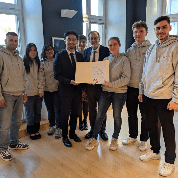 A group of people standing in a room with large windows. Two individuals in the center, dressed in formal attire, hold a certificate or diploma. Others around them wear grey hoodies with 'UE' printed on them.