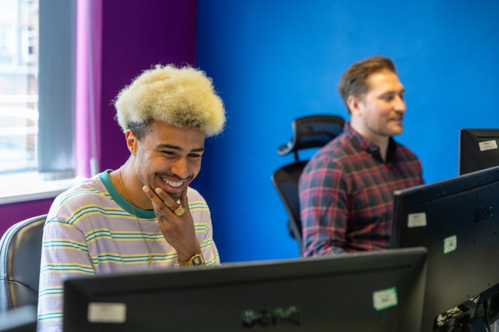 Two men working at desktop computers in a colourful office space. The man in the foreground smiles while looking at his screen, wearing a striped shirt and gold jewellery. The man in the background wears a checkered shirt, with vibrant blue and purple walls adding energy to the workspace.