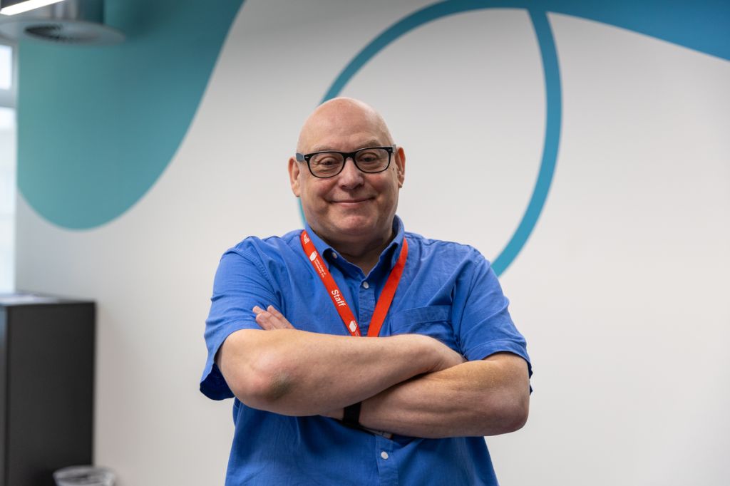 A man in a blue shirt and red lanyard stands confidently with arms crossed, smiling at the camera. He is positioned against a modern office wall with abstract blue designs, creating a professional and approachable impression.