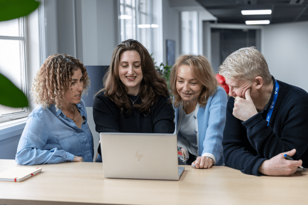 Four colleagues gather around a laptop at a wooden table, smiling and engaged in discussion. The group appears collaborative and focused, with natural light streaming in from nearby windows in a modern office setting.