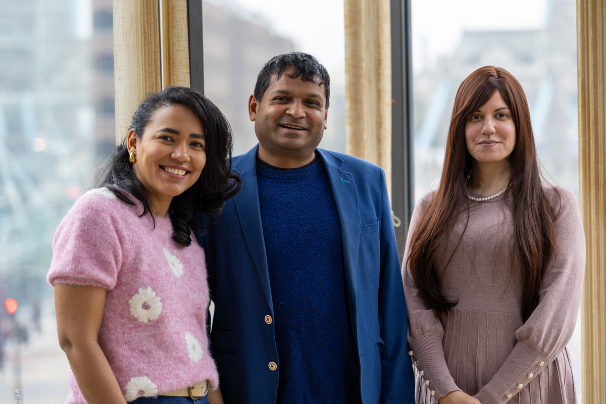 Three colleagues standing together and smiling in front of a window with a city view. The woman on the left wears a pink jumper with white flowers, the man in the middle wears a blue blazer over a matching jumper, and the woman on the right wears a dusty pink dress with a pearl necklace. The backdrop includes glass buildings and a partial view of Tower Bridge, suggesting a London location.