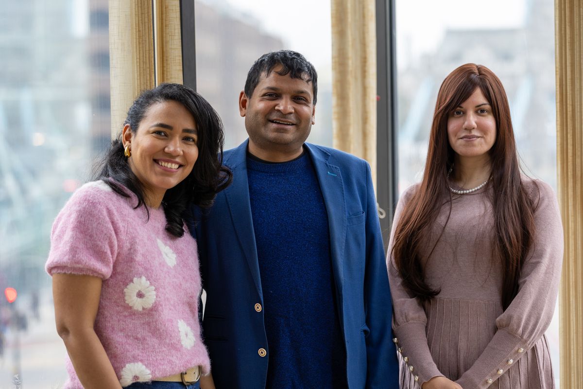 Three colleagues standing together and smiling in front of a window with a city view. The woman on the left wears a pink jumper with white flowers, the man in the middle wears a blue blazer over a matching jumper, and the woman on the right wears a dusty pink dress with a pearl necklace. The backdrop includes glass buildings and a partial view of Tower Bridge, suggesting a London location.