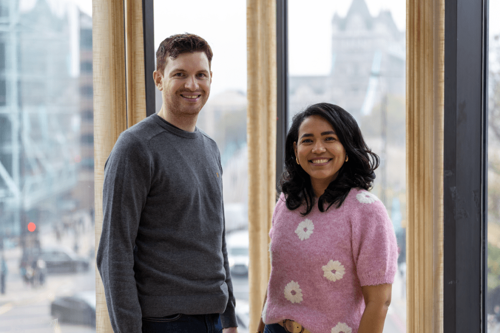 A man and a woman stand smiling in front of large windows with a city view. The man wears a grey jumper, and the woman wears a pink jumper with white flower patterns. Tower Bridge is faintly visible in the background, indicating a London location.