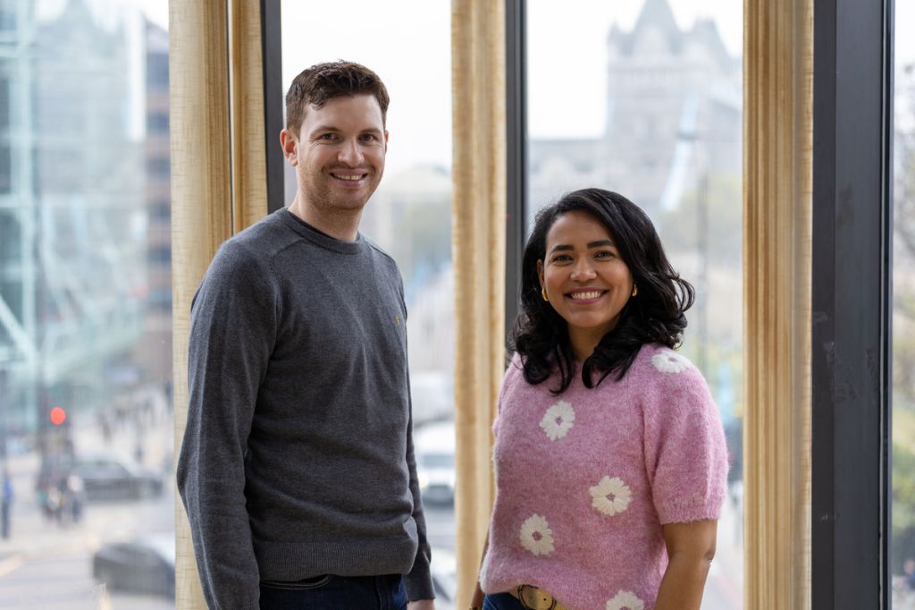 A man and a woman stand smiling in front of large windows with a city view. The man wears a grey jumper, and the woman wears a pink jumper with white flower patterns. Tower Bridge is faintly visible in the background, indicating a London location.
