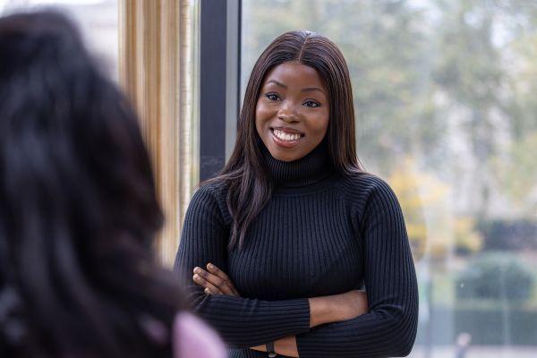 A woman in a black ribbed turtleneck stands near a window with her arms crossed, smiling as she listens to a blurred colleague in the foreground. Natural light fills the space, with soft greenery visible outside.