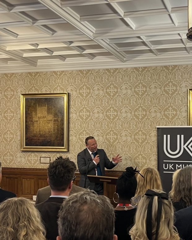 Speaker presenting to seated audience in ornate room with coffered ceiling, traditional paintings, and UK Music banner.