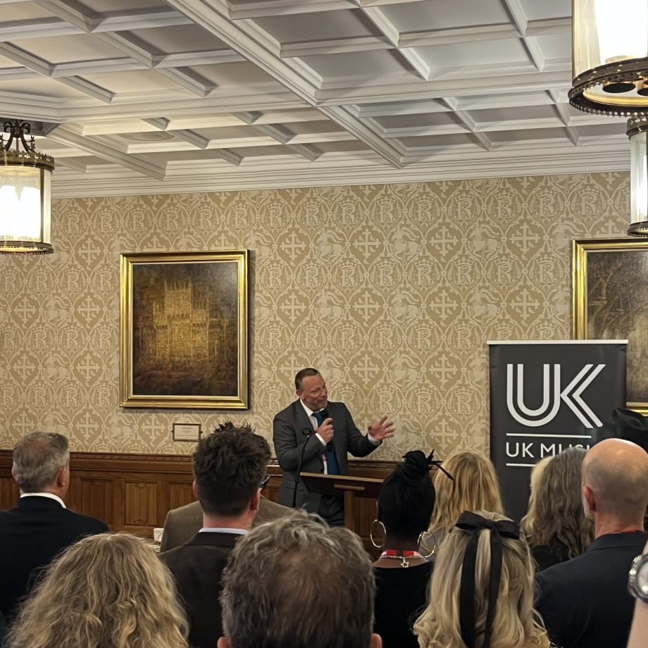 Speaker presenting to seated audience in ornate room with coffered ceiling, traditional paintings, and UK Music banner.