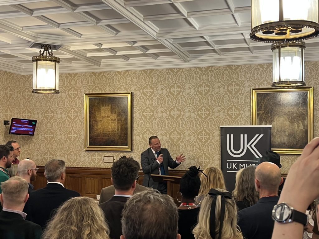 Speaker presenting to seated audience in ornate room with coffered ceiling, traditional paintings, and UK Music banner.