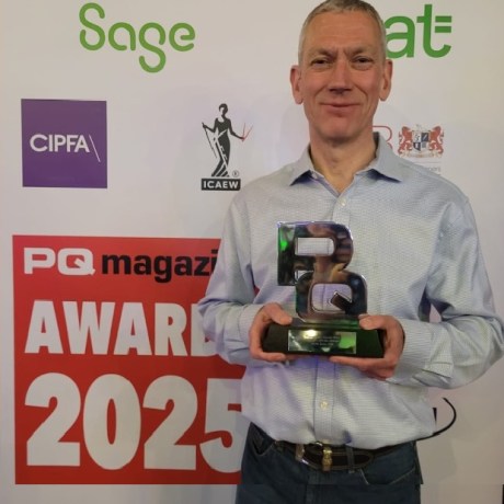 Man in light blue shirt holding a trophy at PQ magazine Awards 2025, standing next to red and black 'WINNER' banner with sponsor logos in background.