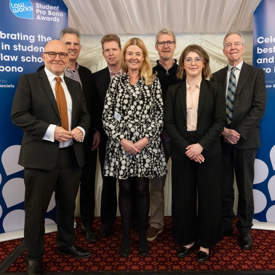 Group of seven people in business attire posing at LawWorks Student Pro Bono Awards event, standing in front of blue branded backdrop.