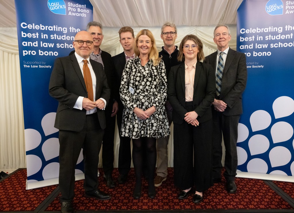 Group of seven people in business attire posing at LawWorks Student Pro Bono Awards event, standing in front of blue branded backdrop.