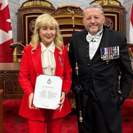 Image of Cyndi McLeod, CEO of GUS Canada and man in formal attire with military medals holding certificate in ornate chamber with Canadian flags.