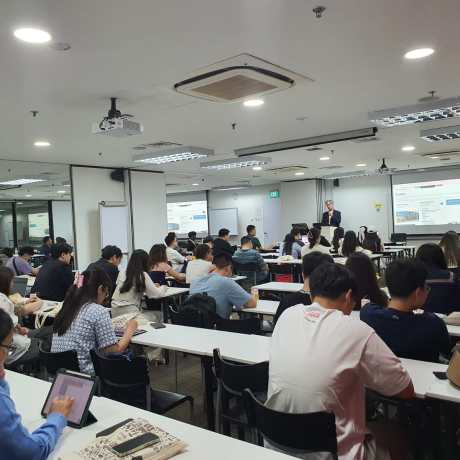 Students seated at desks in a modern classroom listening to an instructor presenting at the front with projected slides.