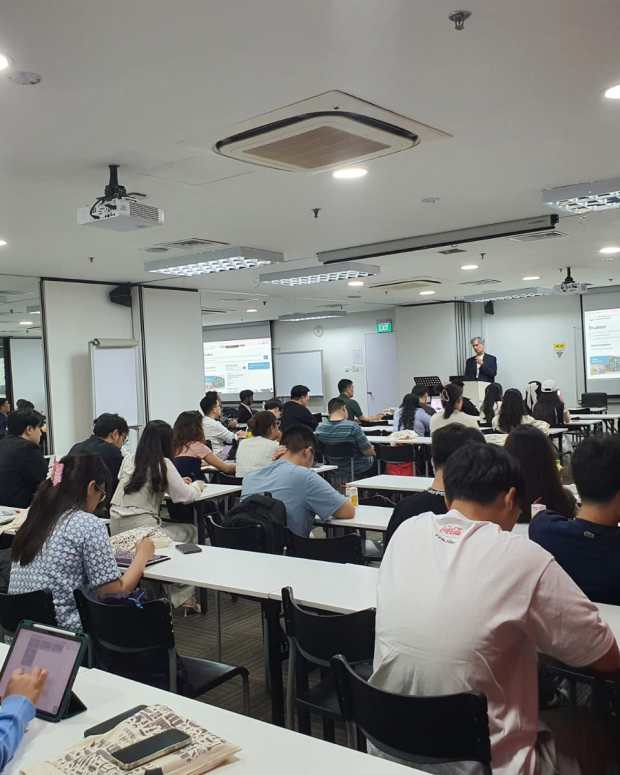Students seated at desks in a modern classroom listening to an instructor presenting at the front with projected slides.