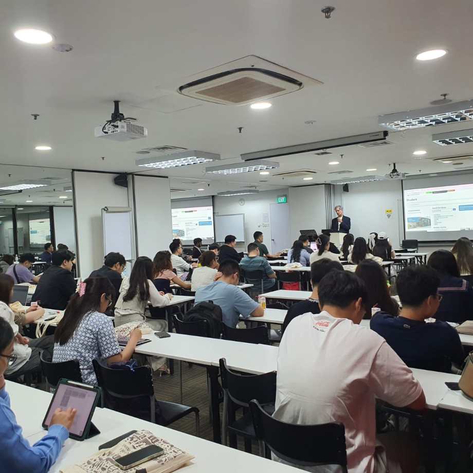 Students seated at desks in a modern classroom listening to an instructor presenting at the front with projected slides.