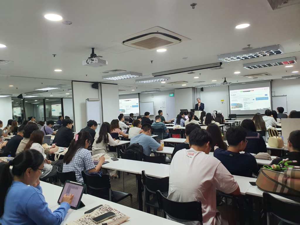Students seated at desks in a modern classroom listening to an instructor presenting at the front with projected slides.