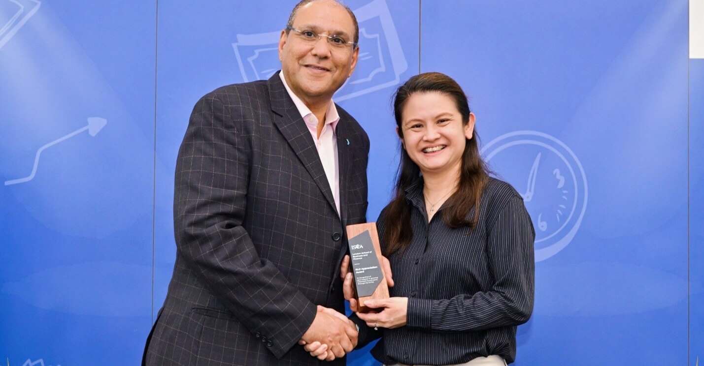 Man in suit and woman in striped shirt shaking hands while holding award against blue backdrop.