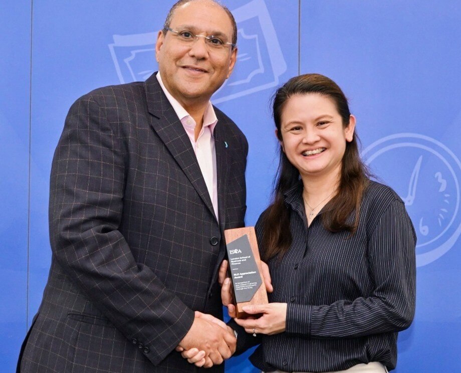 Man in suit and woman in striped shirt shaking hands while holding award against blue backdrop.
