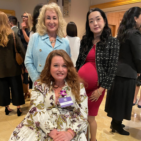 Three women at formal event, one seated in floral dress, two standing in blue and black outfits.