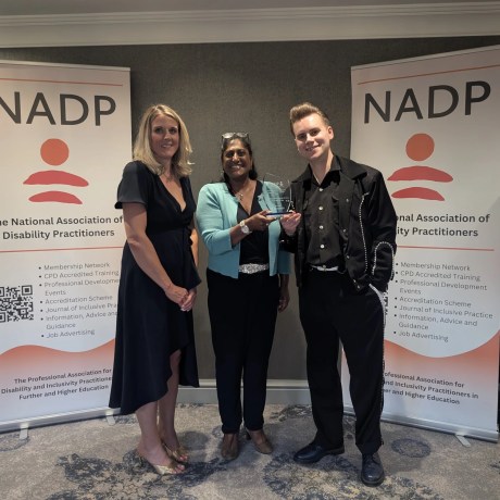 Three people posing with an award at an NADP conference, standing between two organisational banners.